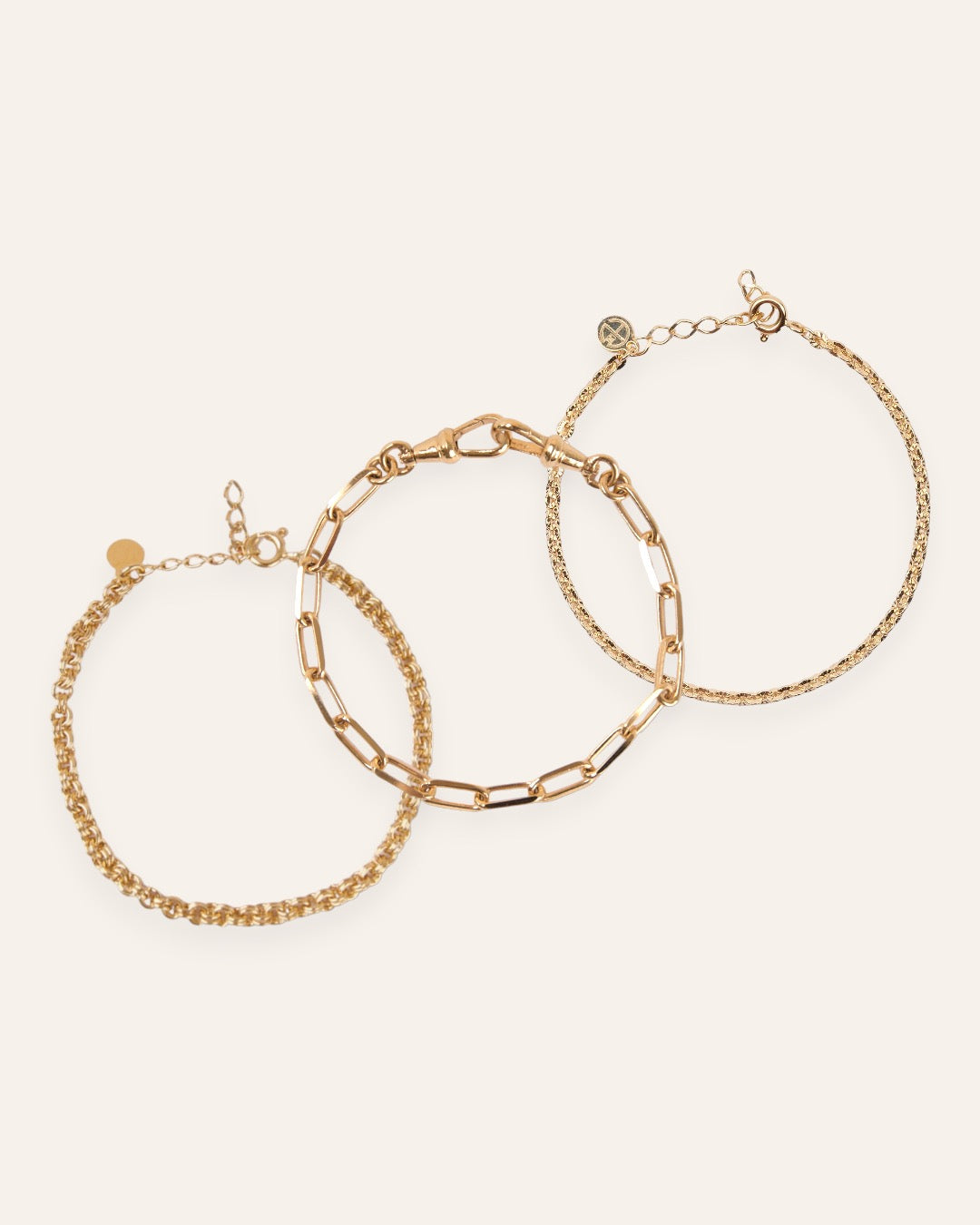 Composition de trois bracelets, avec un bracelet épais en chaîne forçat avec des mailles rondes, un bracelet épais avec une chaîne aux mailles rectangles, et un bracelet en maille soleil en plaqué or 18 carats.