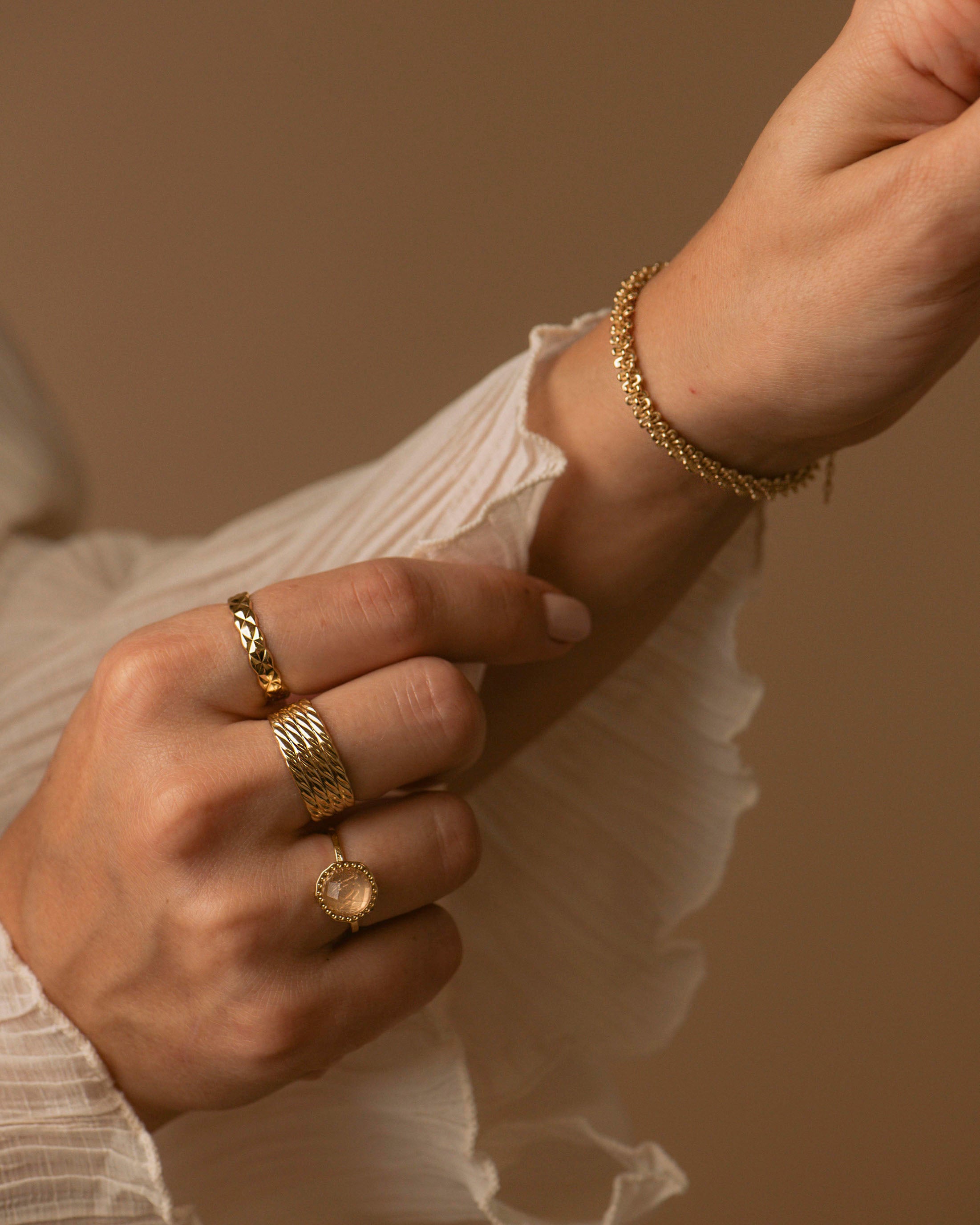 Combinaison de bijoux durables, composée d'une bague fine au motif losange, d'une bague large au motif wavy sur quatre rangées, d'une bague fine ornée d'une pierre de Quartz Rose facettée insérée dans un cabochon, et d'un bracelet en chaîne épaisse avec des mailles entrelacées donnant un effet torsadé, en plaqué or 18 carats made in France.