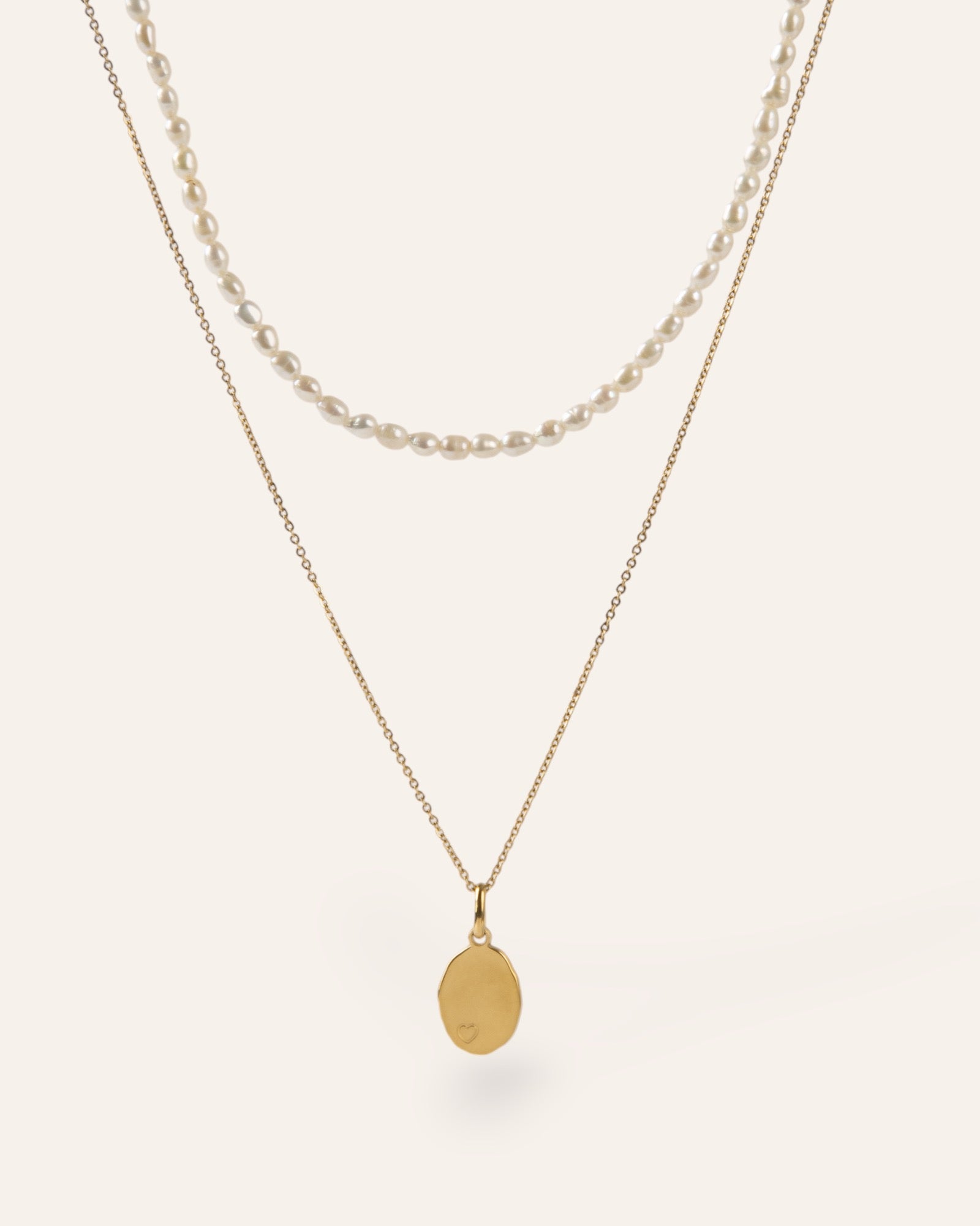 Composition de deux colliers, avec un collier orné de petites perles de culture, et d'un collier en chaîne forçat accompagné d'une médaille de forme ovale avec des contours irréguliers un motif en petit cœur, en plaqué or 18 carats made in France.