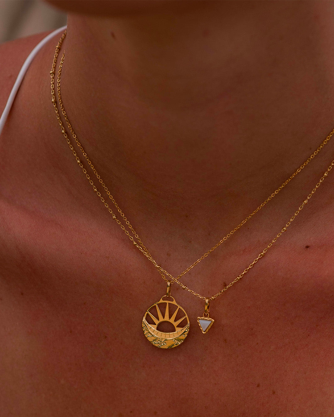Association de colliers éthiques en plaqué or 3 microns composé d'un collier en chaîne forçat avec une médaille au motif soleil et lune et d'un collier en chaîne forçat fin avec un petit pendentif triangle associé à une petite nacre blanche.
