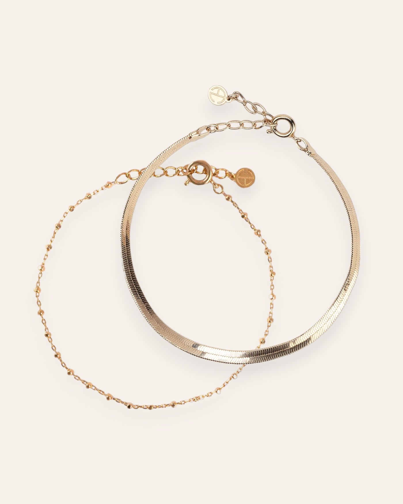Composition de deux bracelets, avec un bracelet en chaîne fine avec des petites mailles carrées, et un bracelet en chaîne plate en plaqué or 18 carats.