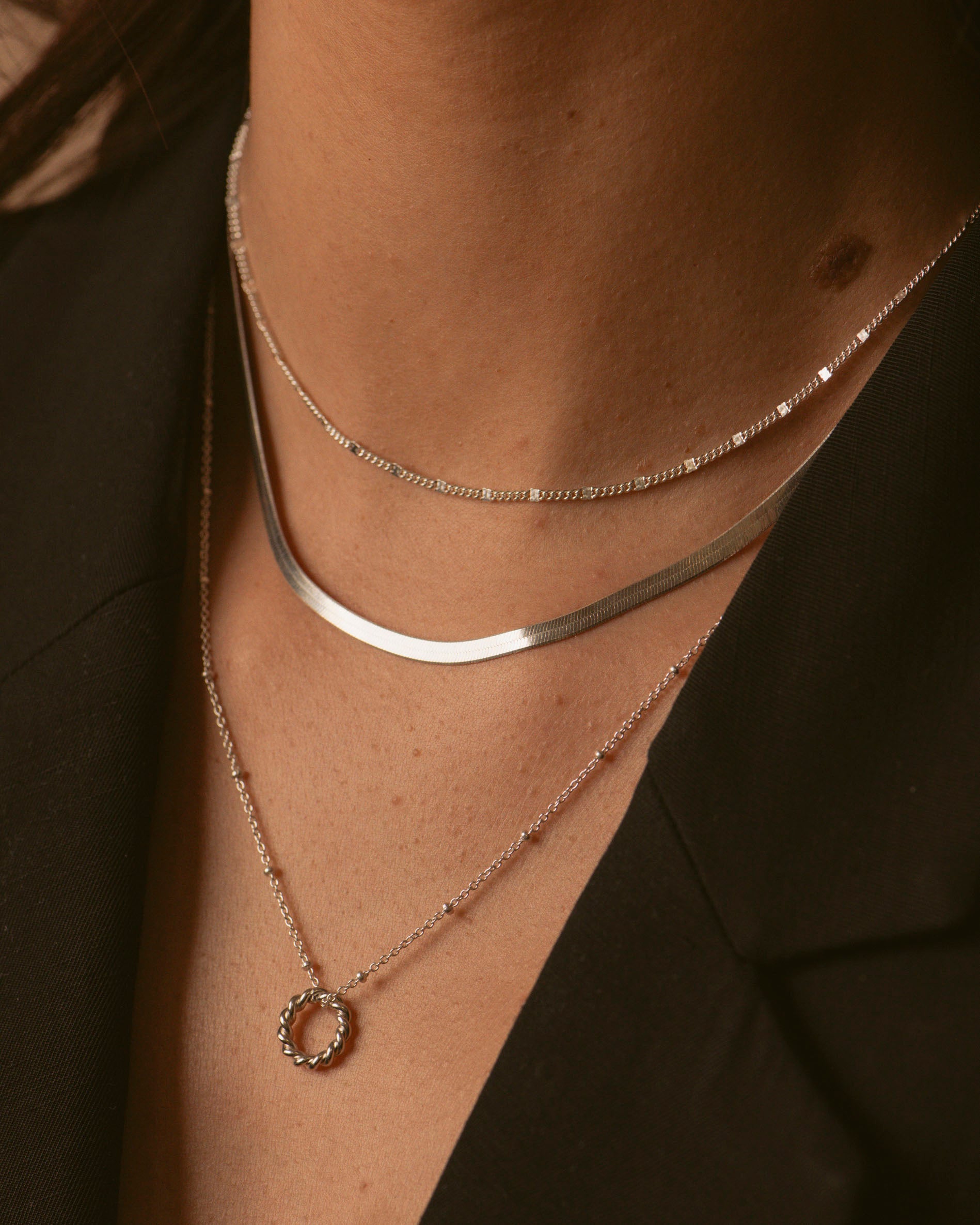 Association de trois colliers en argent massif 925, composée d'un collier ras de cou de type gourmette, d'un collier en chaîne plate, et d'un collier en chaîne boule avec un pendentif en anneau torsadé, made in France.