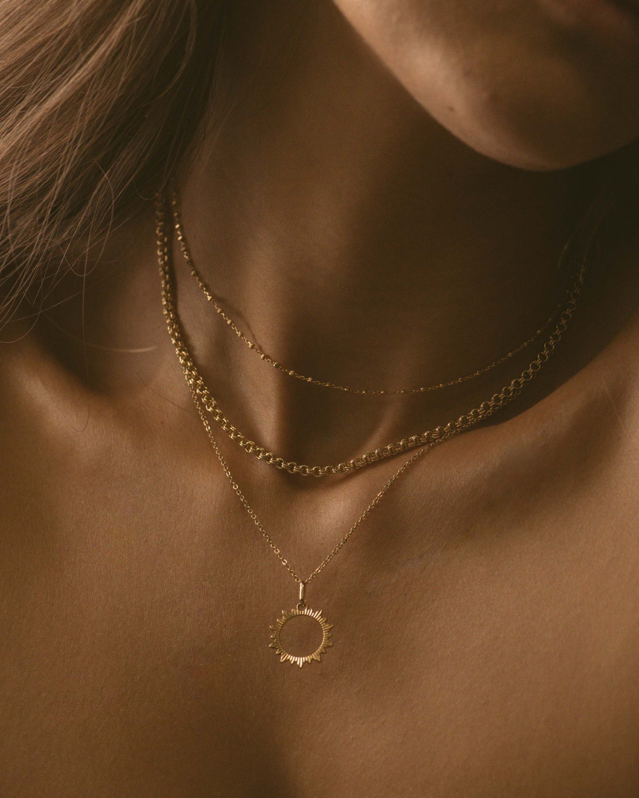 Association de colliers durables en plaqué or 3 microns, composée d'un collier ras-de-cou avec des petites mailles carrées, d'un collier en chaîne forçat avec des mailles rondes épaisses, et d'un collier avec un pendentif soleil ajouré, fabriqués en France.