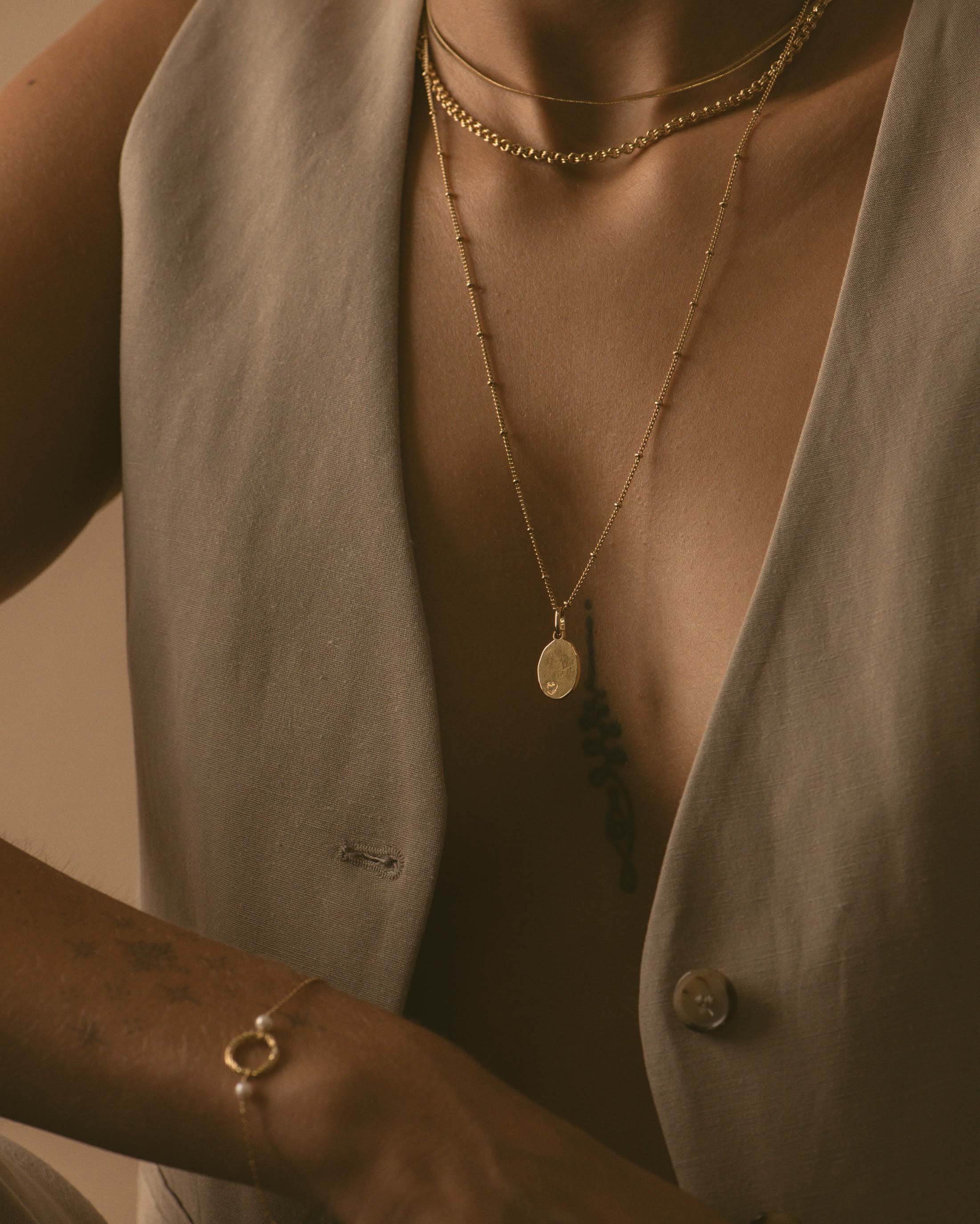 Association de colliers éthiques et responsables, composée d'un collier en chaîne plate, d'un collier ras-de-cou avec des mailles rondes, et d'un sautoir en chaîne boule avec une médaille ovale avec un coeur gravé dessus, et un bracelet composé d'un anneau rayé avec deux petites perles de culture, en plaqué or 3 microns made in France.